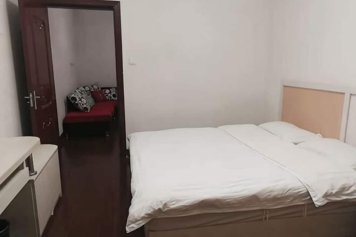 A bedroom with a bed, a red sofa in the background, and a door leading to another room.