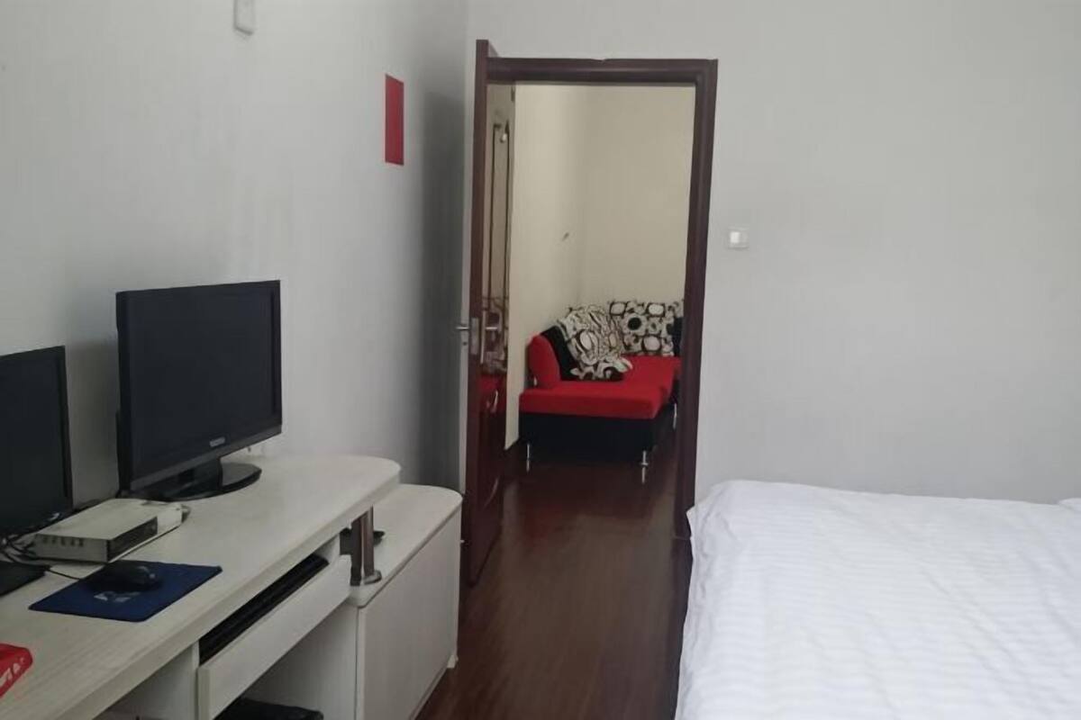 Ein Zimmer mit einem Bett, einem Schreibtisch mit Computer und Bildschirm sowie einer Tür zu einem weiteren Raum mit einer roten Couch.