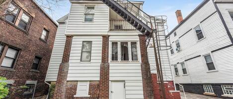 Un edificio de dos pisos con fachada de ladrillo y revestimiento blanco, que cuenta con una puerta central y ventanas, además de una escalera metálica.