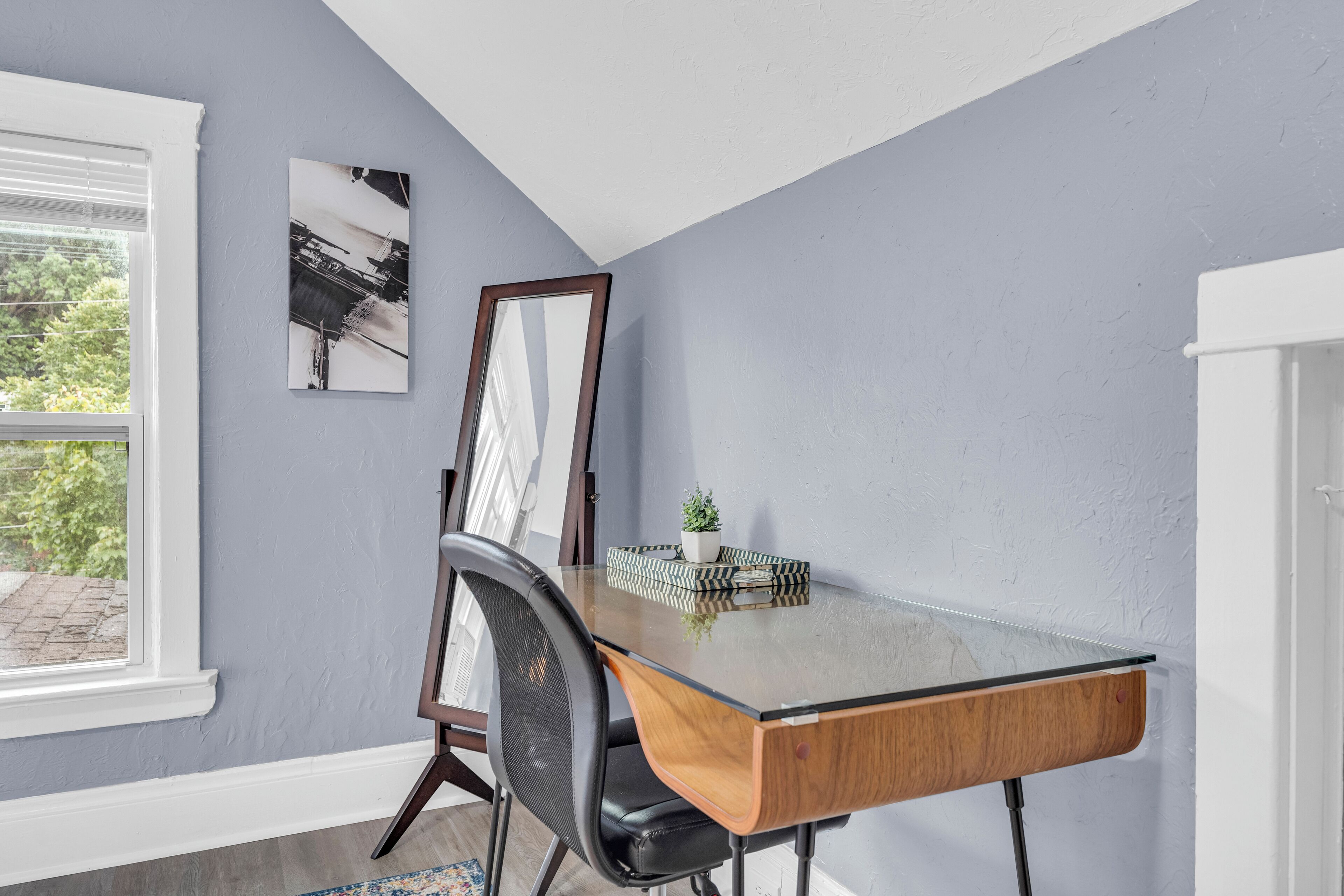 Une pièce comprenant un bureau avec un plateau en verre, une chaise, une petite plante en pot et un tableau encadré accroché au mur.