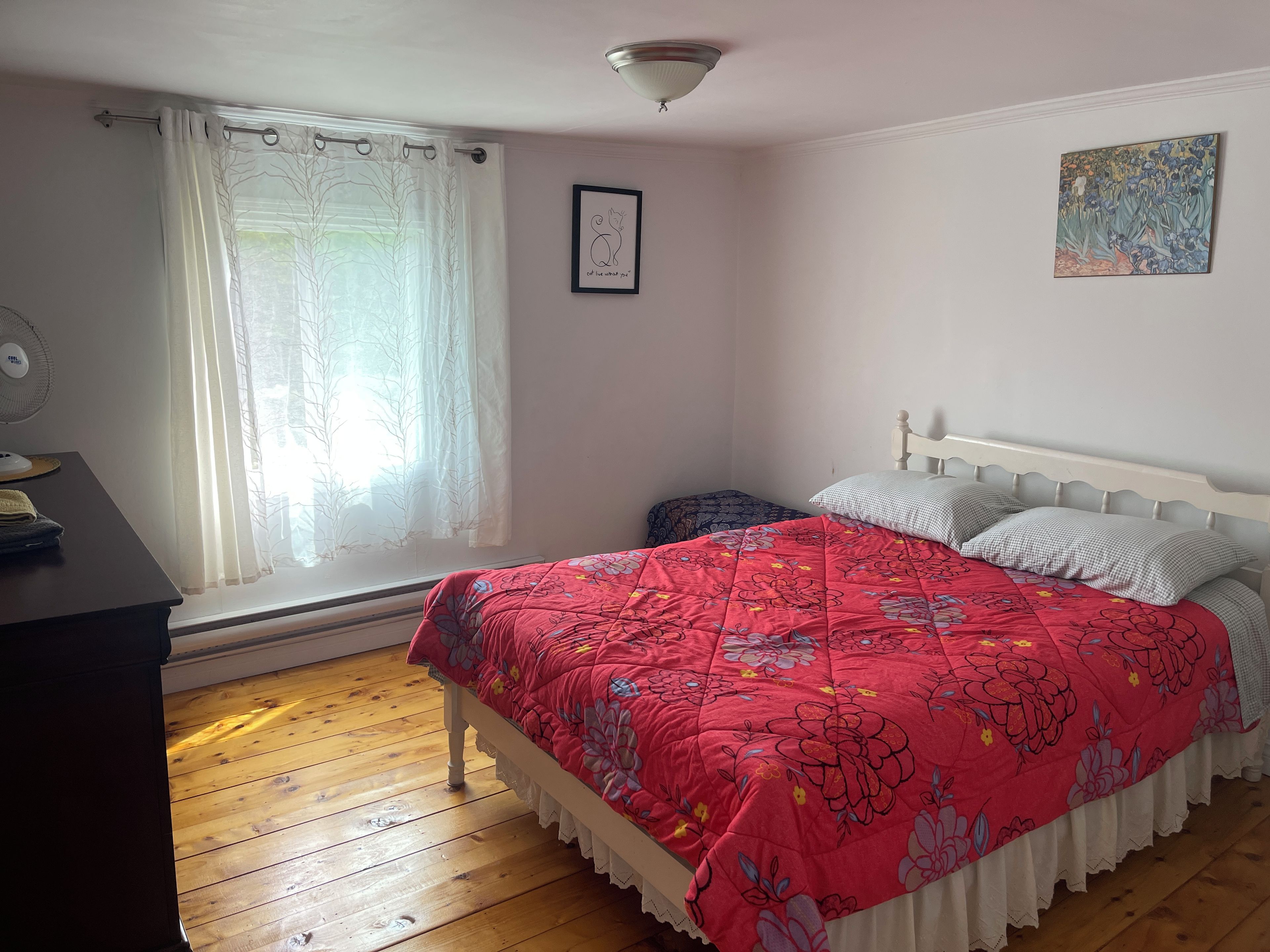 A bedroom with a wooden floor, a bed with a floral bedspread, a white headboard, a bedside table with a lamp, and a window with sheer curtains.