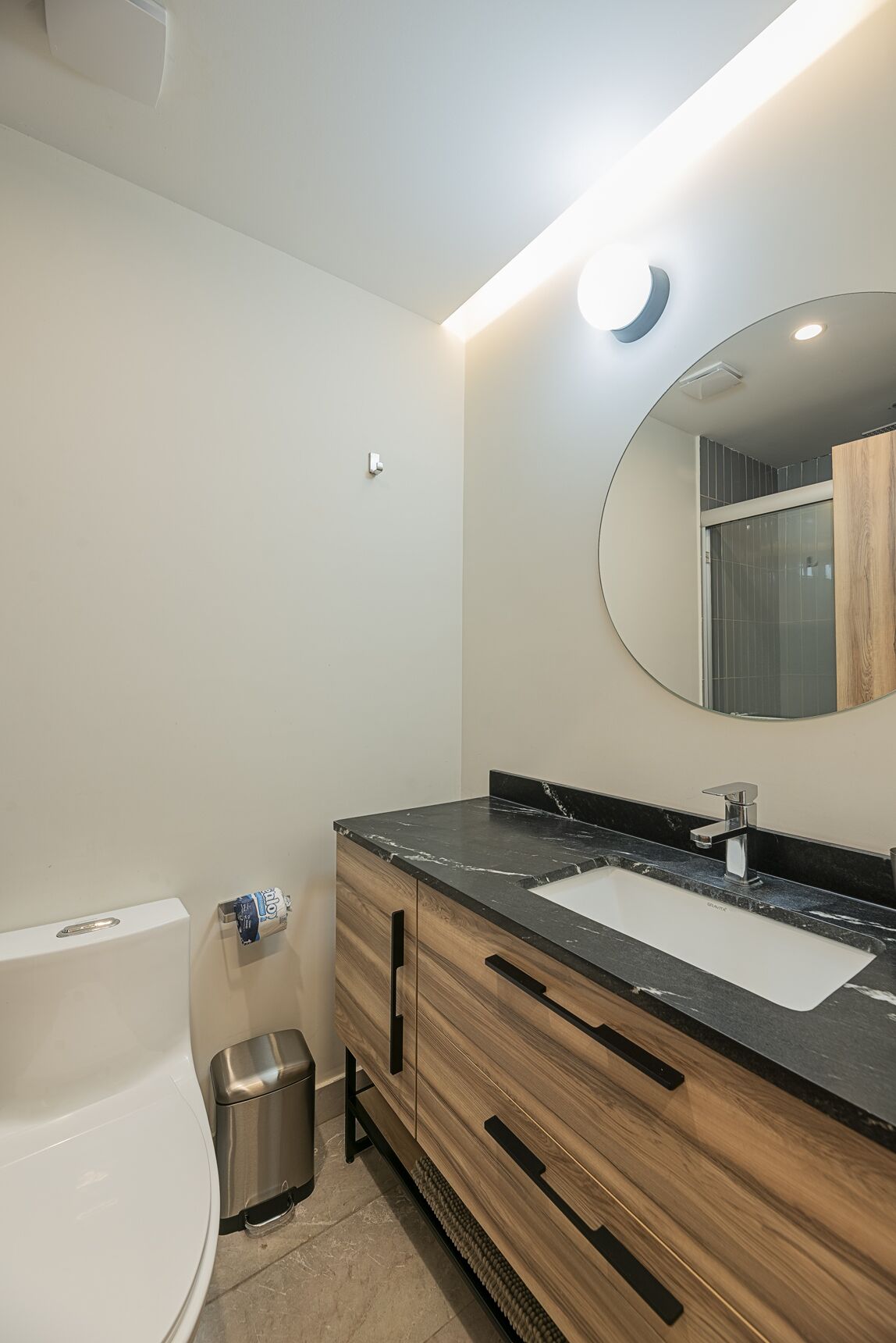 Une salle de bain moderne avec des toilettes blanches, un meuble-lavabo en bois surmonté d’un plan de travail en marbre noir, un grand miroir et un luminaire fixé au mur.