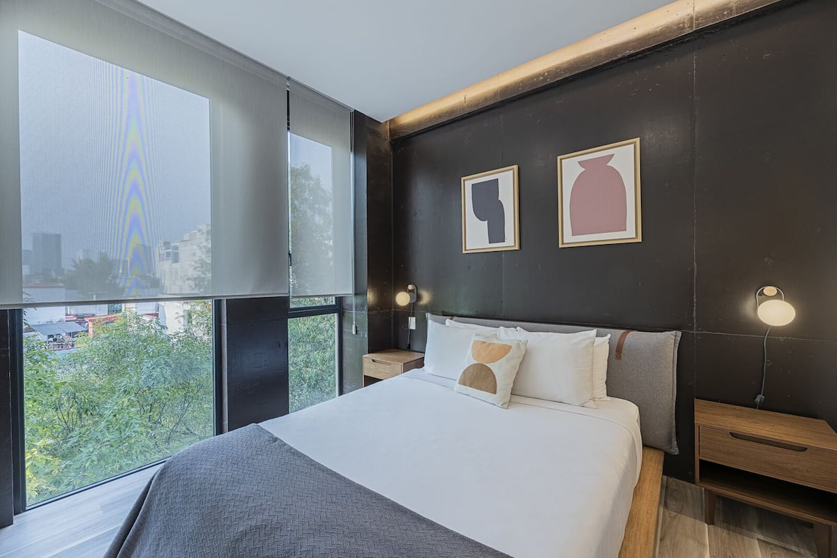 Un dormitorio moderno con una cama grande, dos mesitas de noche y vistas a la ciudad a través de ventanas de gran altura.