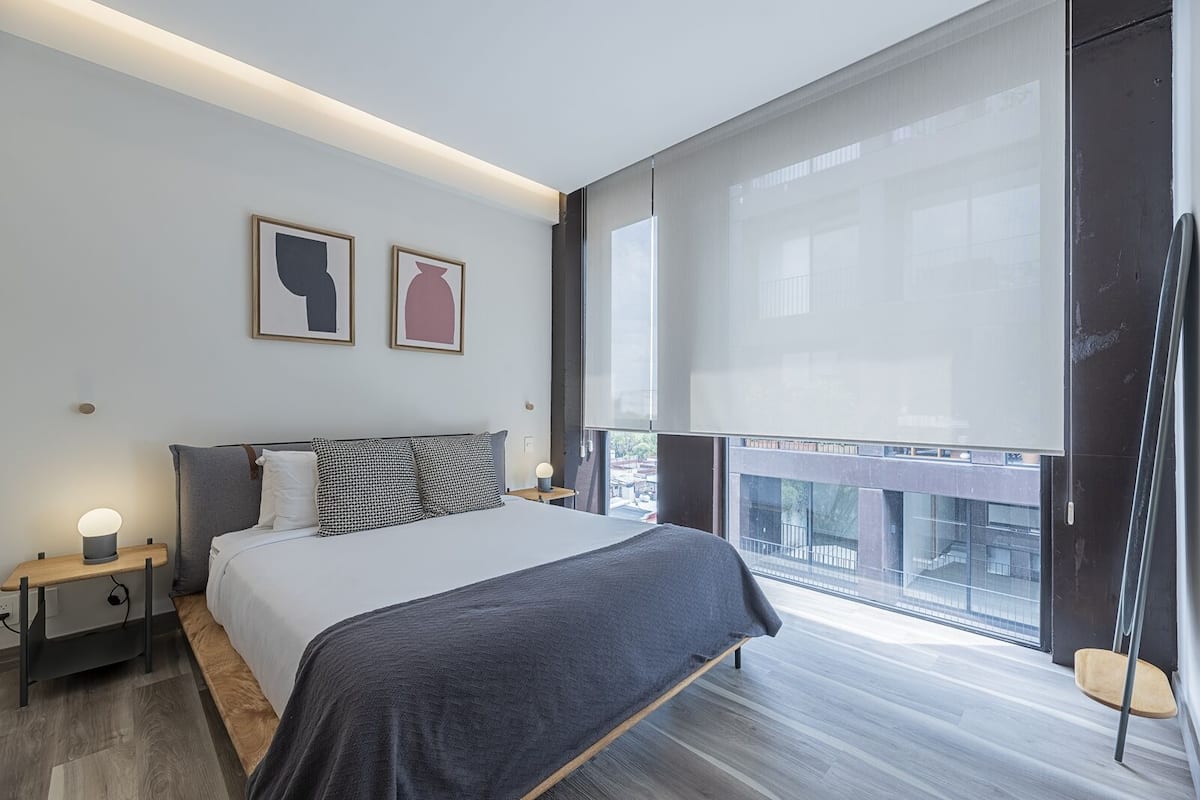 Un dormitorio moderno con una cama grande, mesitas de noche y una ventana con vistas a edificios.