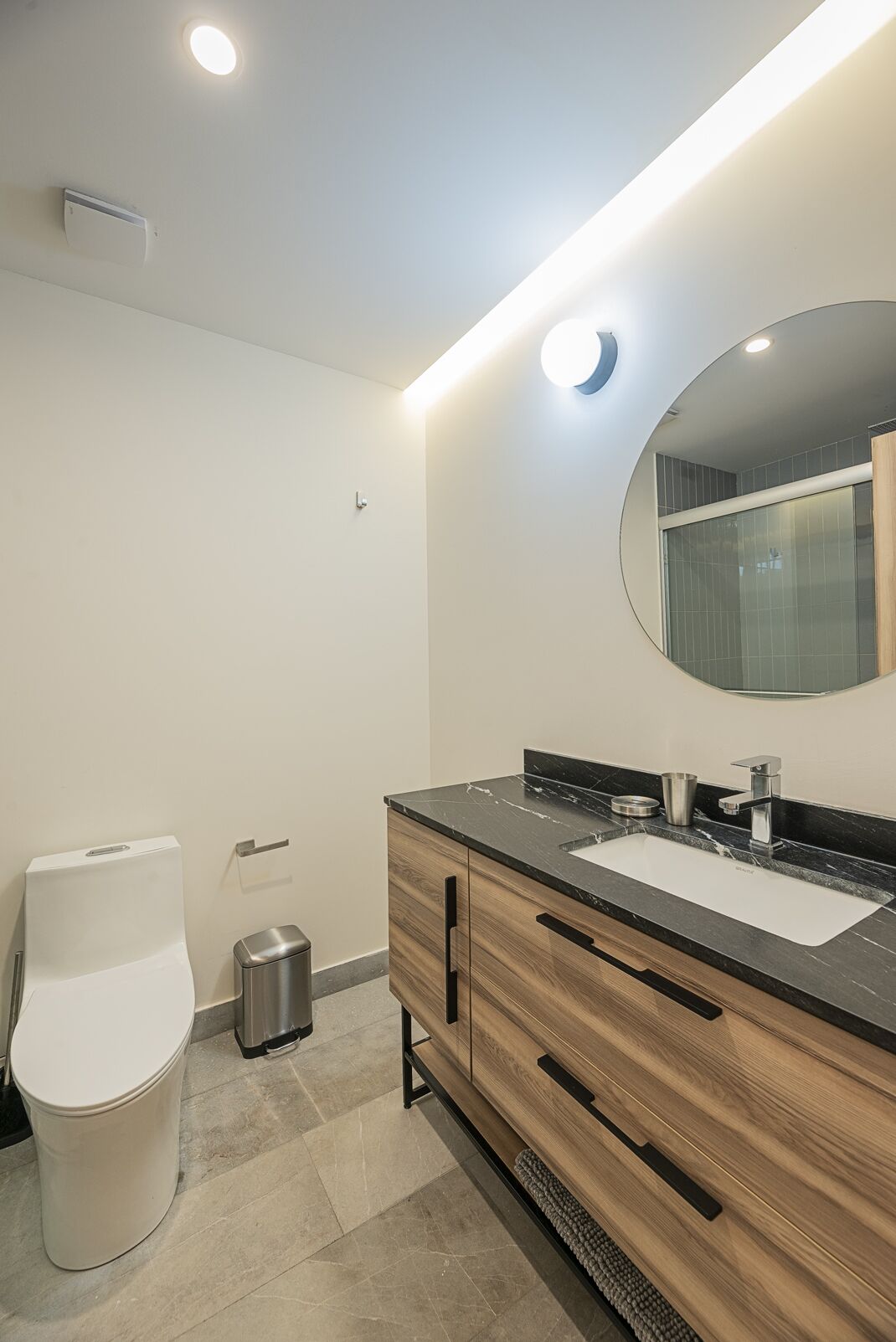 A modern bathroom with a white toilet, a stainless steel trash bin, a wooden vanity with a black countertop, and a large mirror.