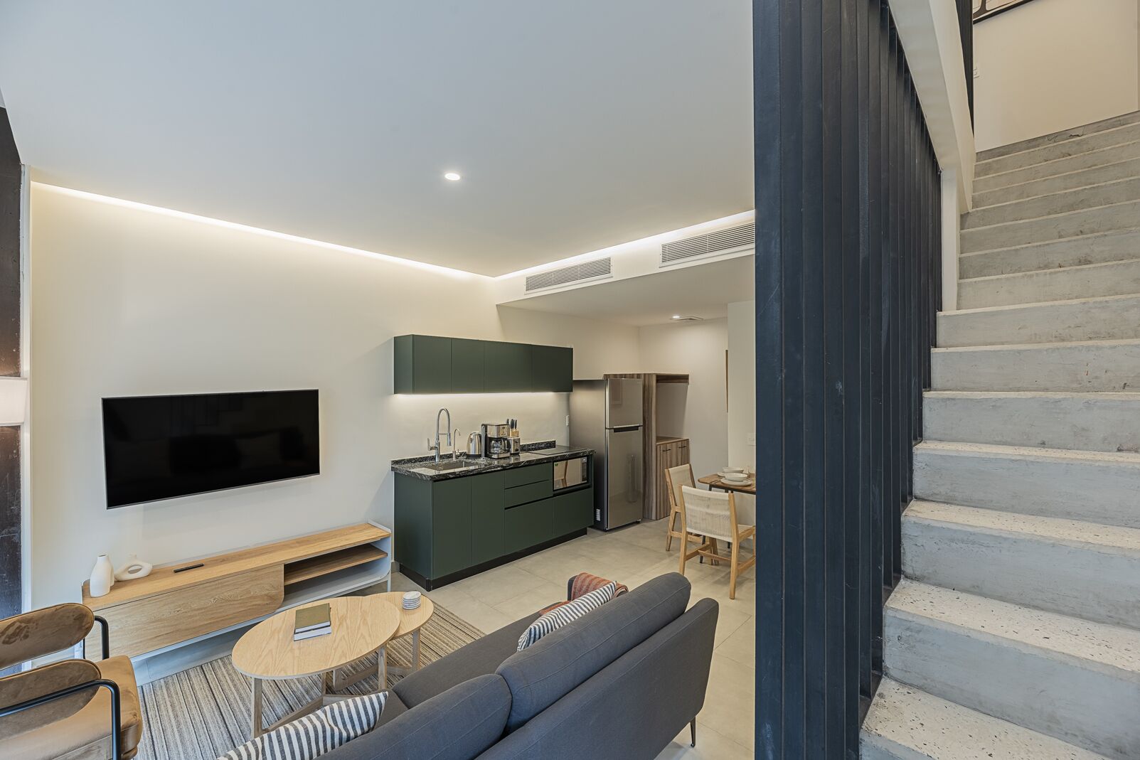 A modern living room with a flat-screen TV, a sofa, a coffee table, and a kitchen area with a refrigerator and cabinets.