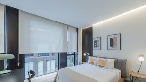 Een moderne hotelkamer met een groot bed, een bureau en een stoel. Er zijn grote ramen en kunstwerken aan de muren.