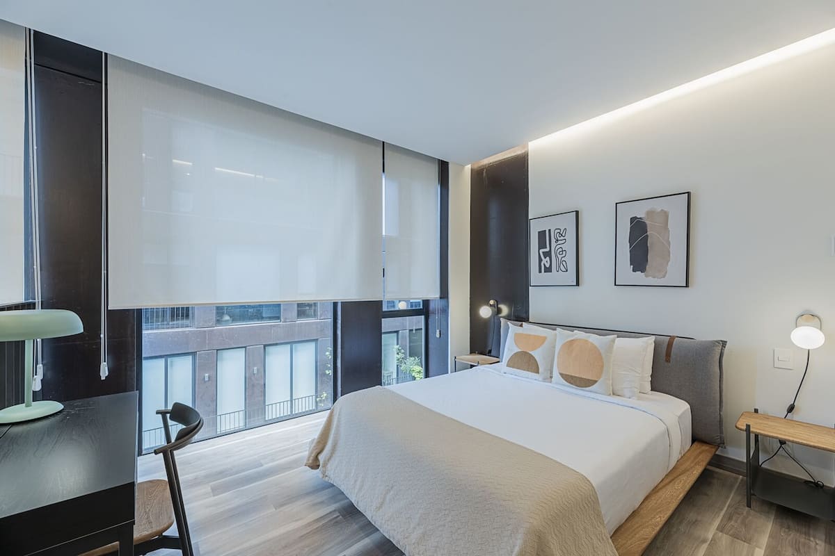 Habitación de hotel moderna con una cama grande, un escritorio y una silla. Cuenta con ventanas amplias y obras de arte en las paredes.
