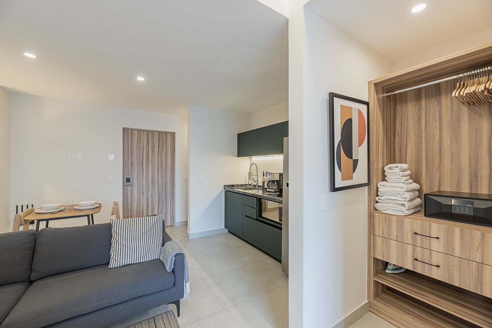 A modern apartment with a grey sofa, a dining table, a kitchen with dark cabinets, and a wooden wardrobe with a towel stack.