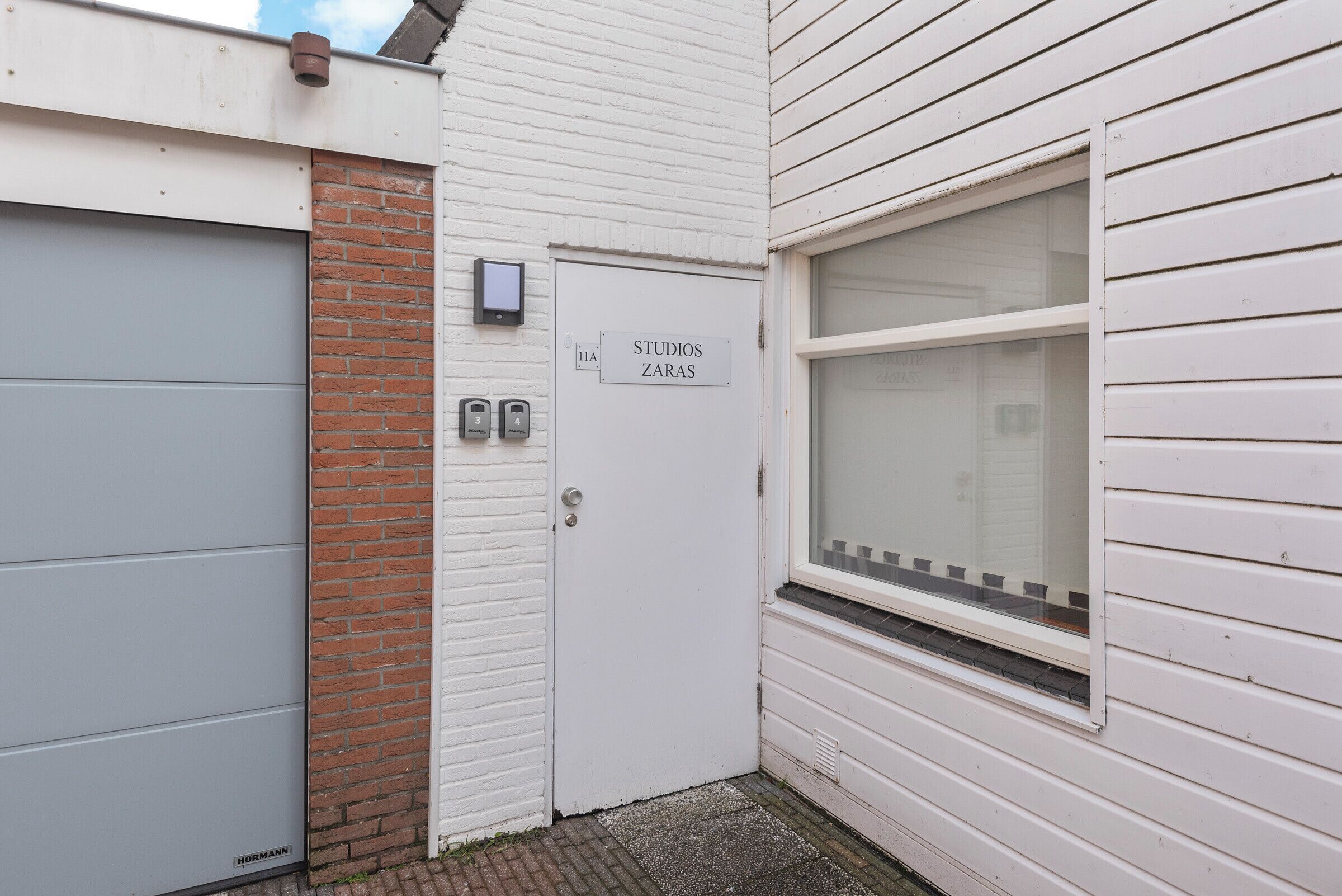 A white building with a closed garage door, a brick wall, and a window with a metal grill.