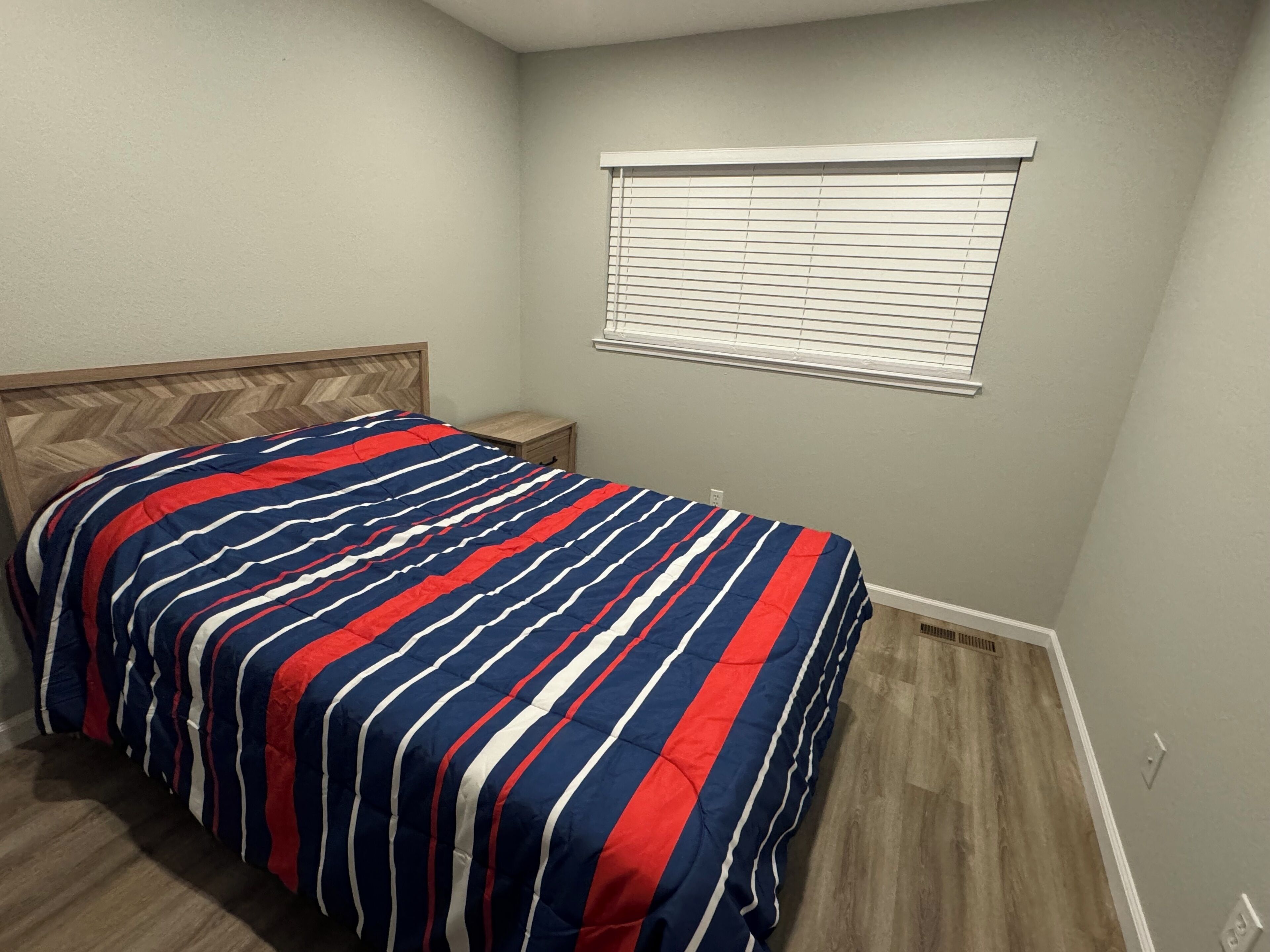 A bedroom with a bed, a nightstand, a window with blinds, and a wooden floor.