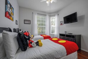 Habitación de hotel con dos camas, un televisor de pantalla plana, un ventilador de techo y peluches de Mickey Mouse.