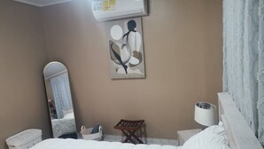 Ein Hotelzimmer mit einem Bett, einer Wandklimaanlage, einem Spiegel, einem kleinen hölzernen Hocker und einer Wandleuchte.