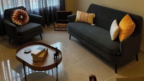 Ein Wohnzimmer mit einem schwarzen Sofa, einem schwarzen Sessel, einem kleinen Holztisch, einem Spiegel an der Wand und einem Fenster mit Vorhängen.