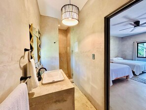A bathroom with a sink, towel rack, and shower. There is a bed visible through an open door.