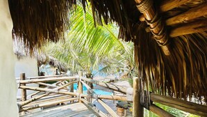 Une terrasse en bois surmontée d’un toit de chaume, une piscine et des palmiers.