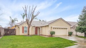 A single-story house with a garage, a well-maintained lawn, and a tree in the front yard. - Fresno House - BBQ, Fire Pit, 3 Bed, 2 Bath & 2 Car Garage! (Fresno)
