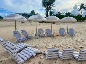 Una playa con sillas de sol a rayas y sombrillas, palmeras y una casa al fondo.