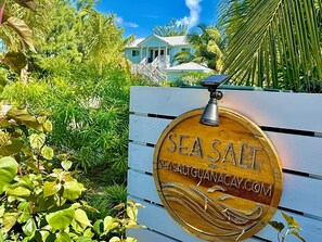 Een houten bord met de tekst 'SEA SALT' en een websiteadres, omringd door weelderige begroeiing en een huis op de achtergrond.