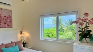 Ein Schlafzimmer mit Bett, Kissen, Nachttisch, Lampe, einem Fenster mit Blick ins Grüne und einer Zimmerpflanze.