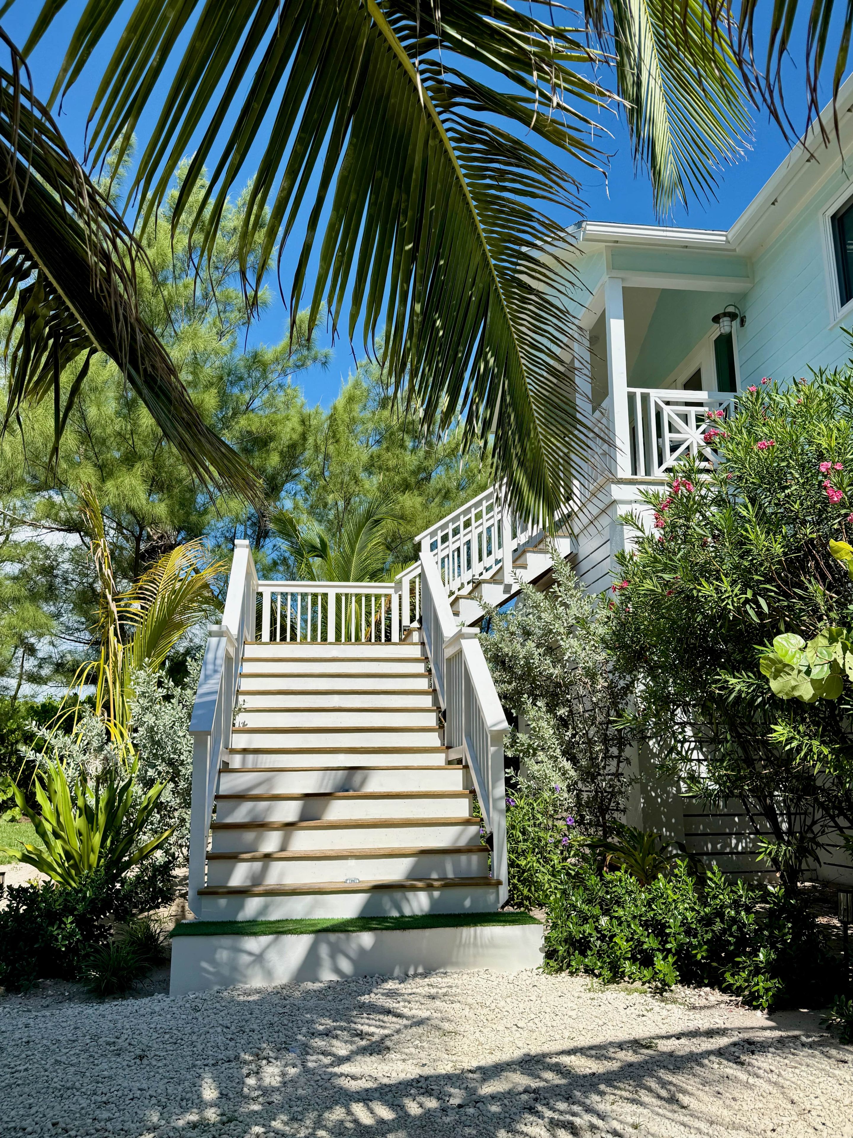 A staircase leading to a house with a white railing, surrounded by greenery and a clear blue sky.