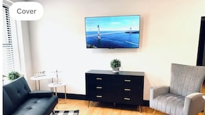 Ein modernes Wohnzimmer mit einer schwarzen Couch, einem grauen Sessel, einer schwarzen TV-Konsole mit einer Pflanze und einem karierten Teppich.