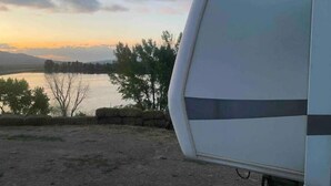 A white RV parked near a body of water with trees and a stone wall in the background. - Lake shore queen bed trailer, tiki torches, & tent space for extra guest (Clifton)