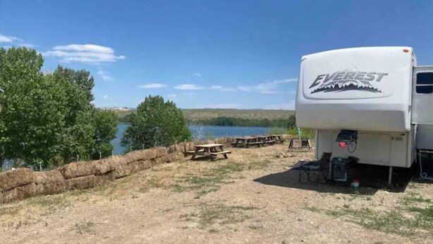 A white RV with 'EVEREST' branding parked near a lake, surrounded by picnic tables and hay bales. - Lake shore queen bed trailer, tiki torches, & tent space for extra guest (Clifton)