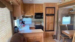 A compact RV kitchen with wooden cabinets, a microwave, stove, and a dining area with a table and chairs. - Lake shore queen bed trailer, tiki torches, & tent space for extra guest (Clifton)