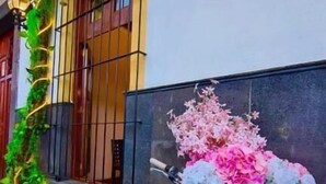 A bicycle with a floral arrangement attached to its frame, parked on a tiled pavement in front of a building with a barred window and wall-mounted lights. - Hotel Boutique Pedregal (Coatepec)
