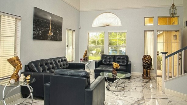 A spacious living room with black leather sofas, a glass coffee table, and a marble floor. There are two windows with blinds, a large painting of the Eiffel Tower, and a staircase with a railing. - Charming 3-bedroom house in Moreno Valley with huge pool ,spa and connected lake (Moreno Valley)