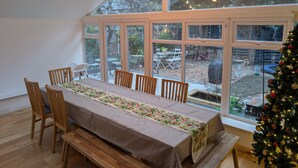 A dining area with a long table, wooden chairs, and benches. It features large windows with a view of the outdoors, including a patio and a Christmas tree. - Cosy Family House | Quiet | Garden | Free Parking (London)
