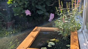 A wooden planter box with a small pond, surrounded by greenery and pink flowers. - Cosy Family House | Quiet | Garden | Free Parking (London)