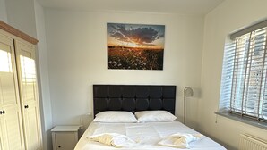 A bedroom with a bed, a bedside table with a book, a window with blinds, a wardrobe, and a wall-mounted picture. - Cosy Family House | Quiet | Garden | Free Parking (London)