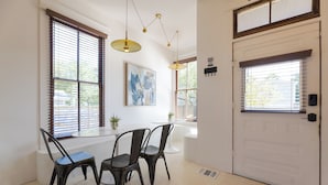 A dining area with a white table, black chairs, and pendant lights. There are windows with blinds and a door with a handle.