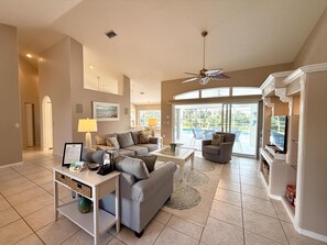 A spacious living room with a grey sofa, a coffee table, and a TV. There is a ceiling fan, a lamp, and a view of the outdoors through large windows.
