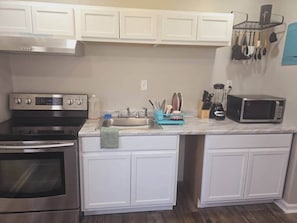 A compact kitchen with white cabinets, a stainless steel oven, a microwave, and a sink with a dish rack. - Comfy 1BR Apt. in Pleasant Ridge (Cincinnati)