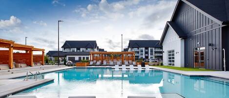 Une piscine entourée de chaises longues, une pergola en bois et, au loin, des bâtiments modernes.
