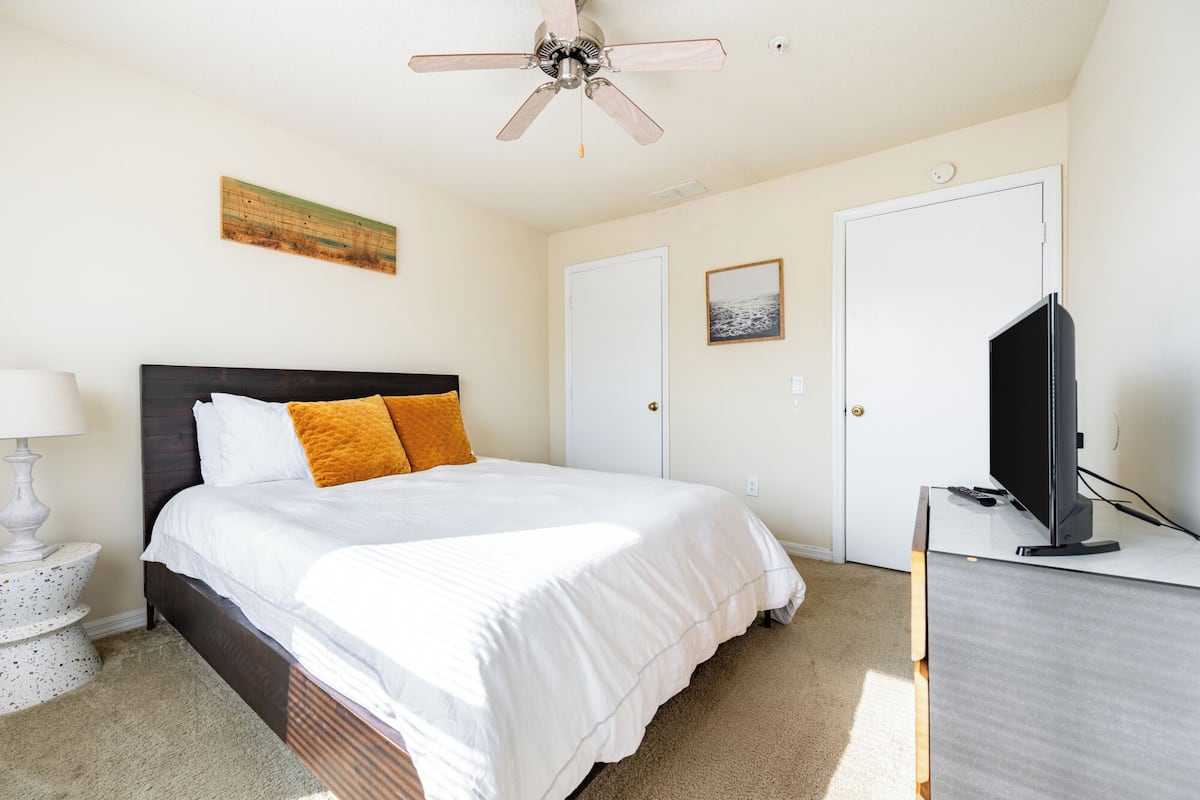 A bedroom with a bed, a ceiling fan, a TV, a desk, and a painting on the wall.