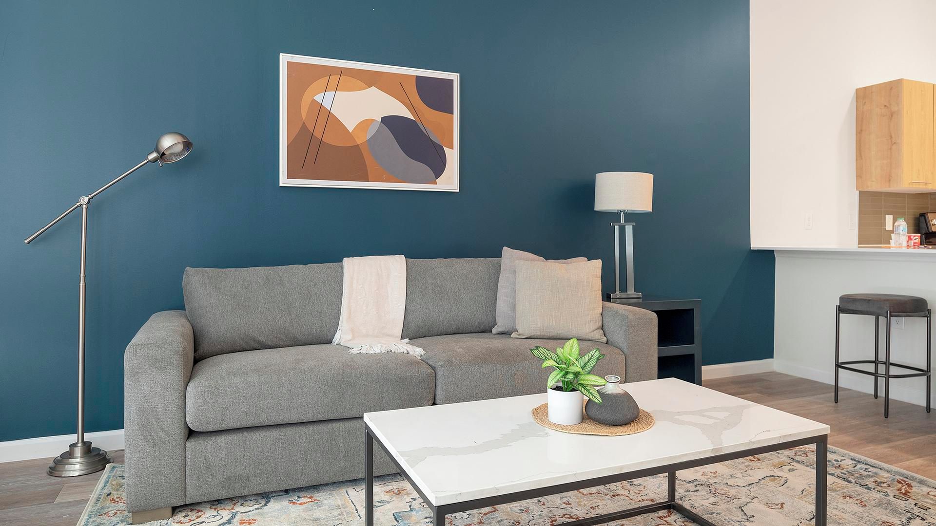 Una sala de estar con un sofá gris, una mesa de centro blanca, una lámpara de pie y un cuadro en la pared.
