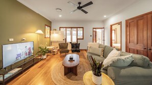 A spacious living room with a flat-screen TV, wooden flooring, a ceiling fan, and a large window with wooden frames. - House of Fairy Wren - Fully renovated charming Queenslander! 10 Mins to BNE CBD! (Annerley)