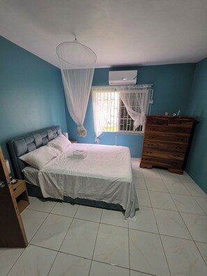 A bedroom with a bed, a dresser, a window with curtains, and an air conditioning unit.