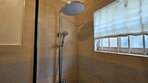 A shower with a rain showerhead and handheld showerhead, a window with blinds, and tiled walls.