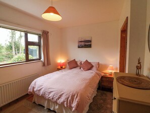 A bedroom with a bed, bedside lamps, a window, a radiator, and a small table with a decorative item. - KNOCKBRACK, pet friendly, with open fire in Killorglin, County Kerry (.)
