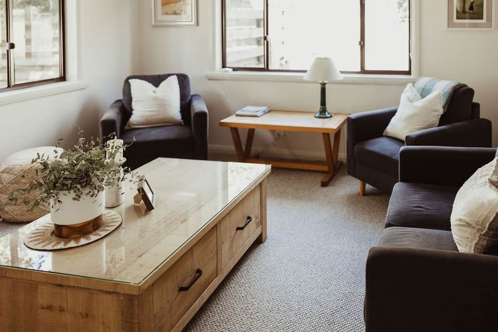 Un salon avec un canapé, une table basse et une lampe. Il y a des fenêtres avec des stores, une plante sur la table basse et un cadre photo accroché au mur.