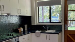 A modern kitchen with white cabinets, a stainless steel range hood, a built-in oven, a black countertop, and a window with a view of greenery. - The Rosella - One-bedroom cottage at Bundaleera Vineyard (Relbia)