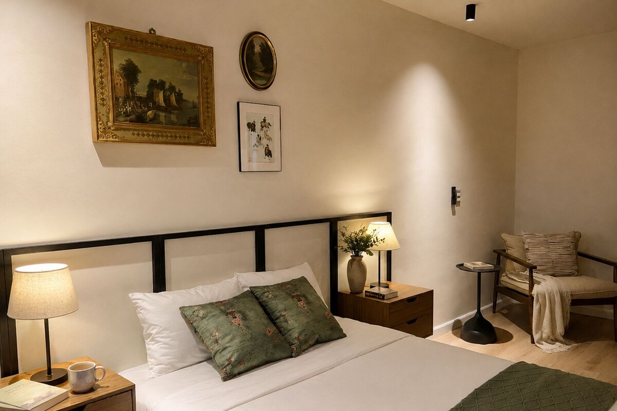 Habitació d'hotel ben ordenada amb un llit, taules de nit, una cadira i obra d'art penjada a la paret.