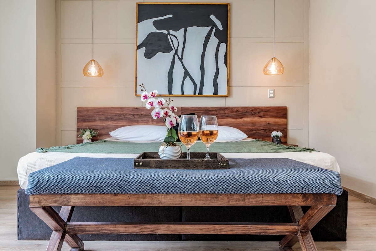 Una cama bien hecha con cabecera de madera, dos lámparas colgantes, una bandeja con dos copas de vino y un arreglo floral sobre la cama.