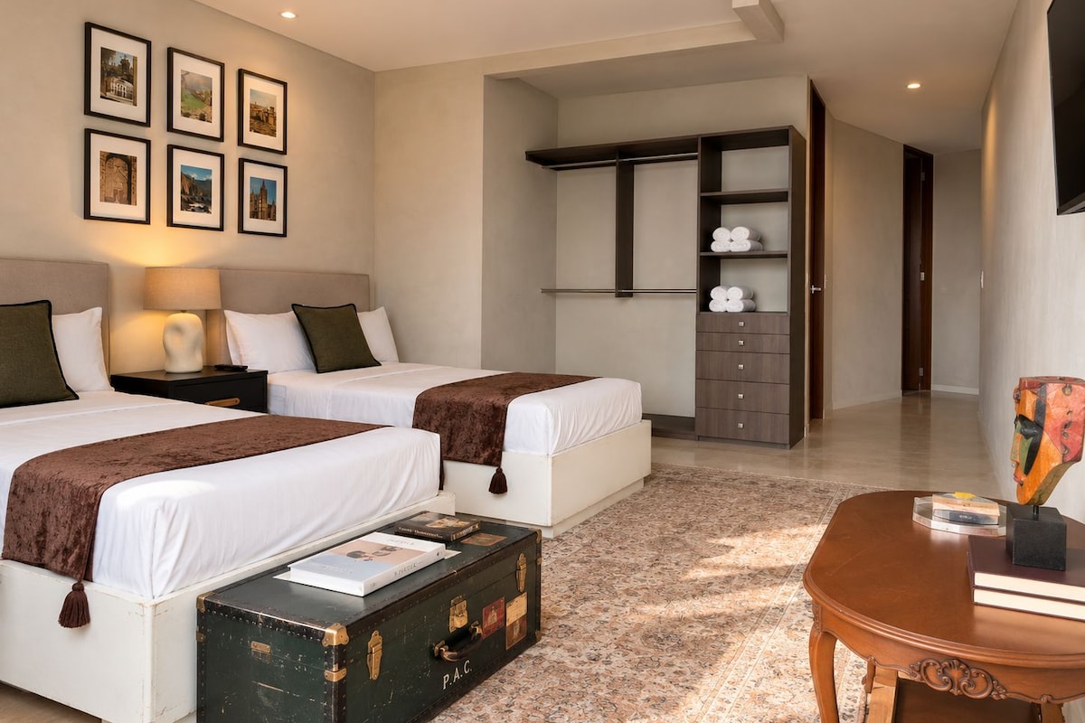 Habitación de hotel con dos camas, un armario, un escritorio y una mesita con libros.