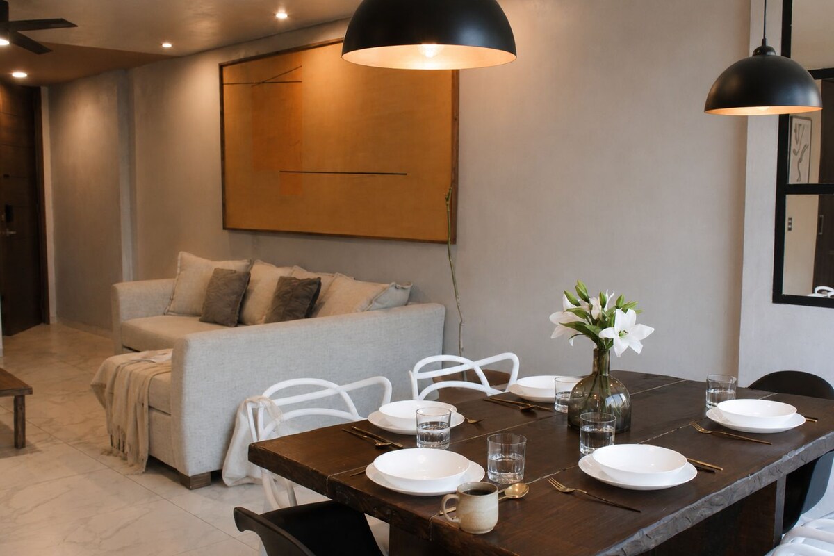 Un comedor con una mesa dispuesta para cuatro personas, un sofá e iluminación colgante moderna.